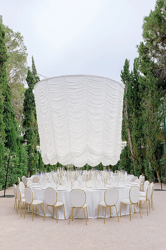 Reception tablescape with white linen round table, glassware and tall taper candles under a draped canopy in an evergreen garden with string lights