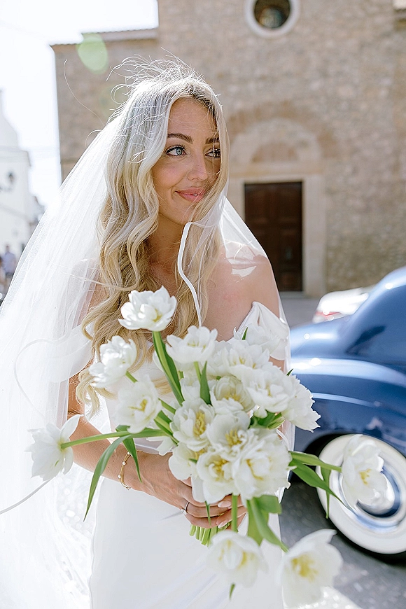Bridal portrait of a bride holding bouquet with a flowing wedding veil, off-shoulder dress, by a stone building and vintage car outdoors