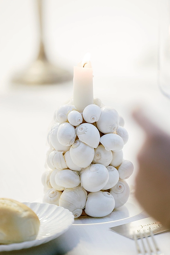 Wedding table centerpiece of seashells surrounding a white taper candle with place card, plates and fork on a white tablecloth in soft daylight