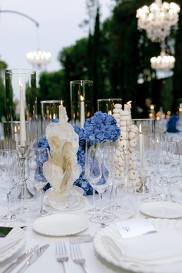 Reception tablescape with blue hydrangea centerpiece, tall taper candles, crystal glasses and macarons under garden chandeliers at dusk