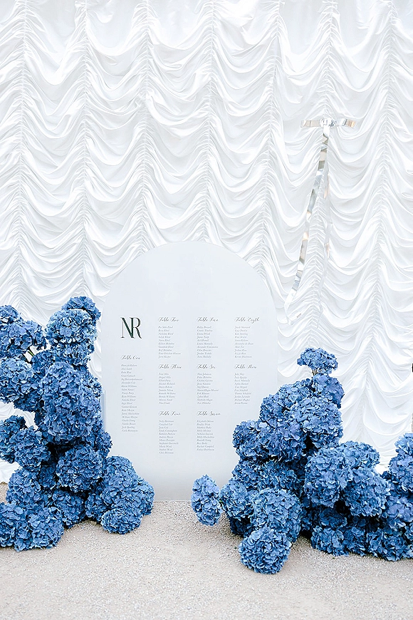 Wedding seating chart with an arched seating chart sign and printed guest list, framed by blue hydrangeas against a white draped backdrop