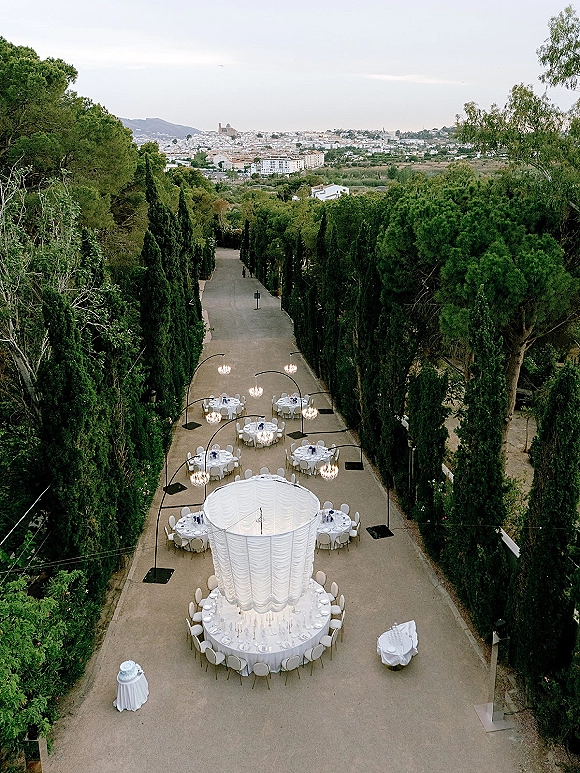 Outdoor reception setup with round tables, white linens, and chandeliers beneath a sheer draped canopy along a tree-lined driveway