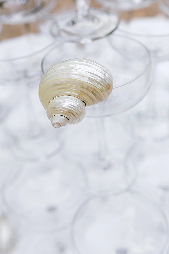 Wedding shell place cards in seashell place card holder beside clear stemware on a crisp white table setting at a coastal reception