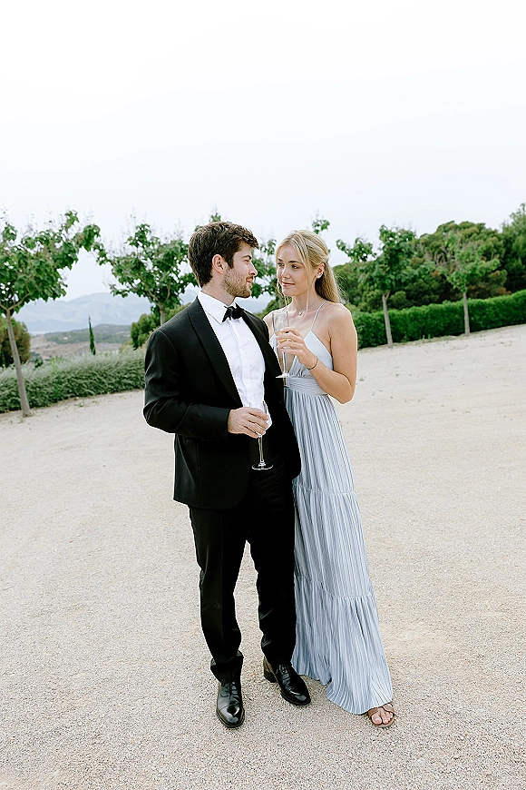 Wedding guest portrait of a couple in black tie formal wedding guest attire holding champagne flutes on a gravel path with trees and hills behind