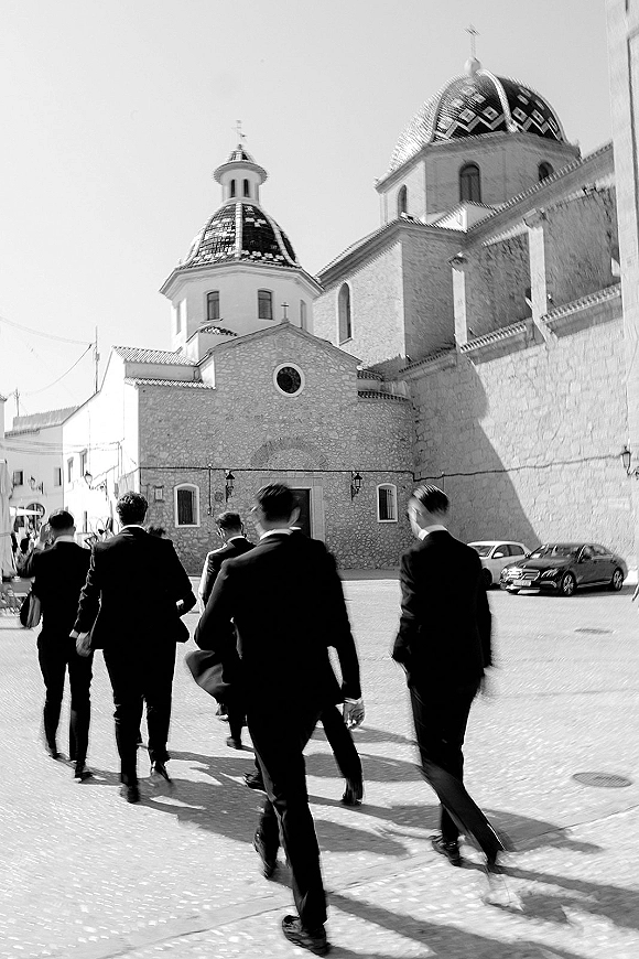 Groomsmen arrival as men in black suits walk up a cobblestone street toward a domed stone church exterior in daylight