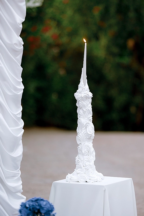 Wedding unity candle with carved wax and steady flame beside a white taper candle on a white tablecloth, set outdoors near greenery