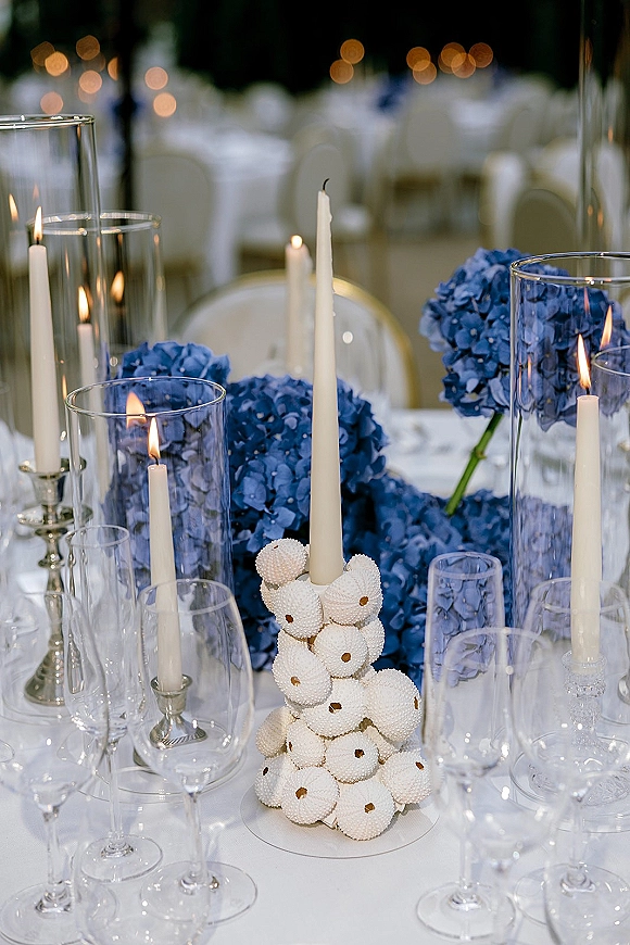 Reception tablescape with wedding table centerpiece of white taper candles in glass hurricane vases and blue hydrangeas, amid soft reception lights