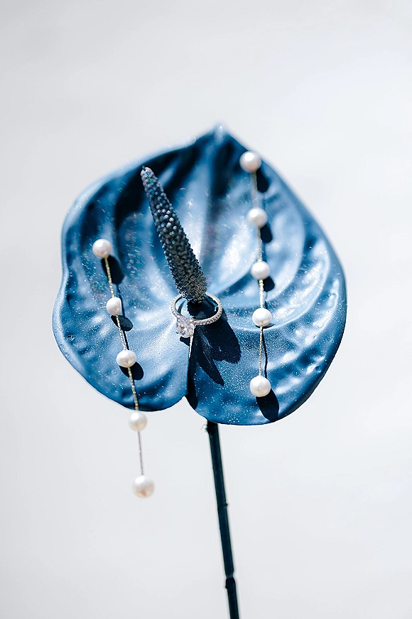 Engagement ring photo of a diamond solitaire on a blue anthurium, styled with a pearl strand against a light neutral backdrop