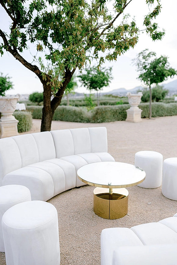 Wedding lounge seating with a white curved sofa, ottomans, and round coffee table with gold base on a gravel garden patio with hedges