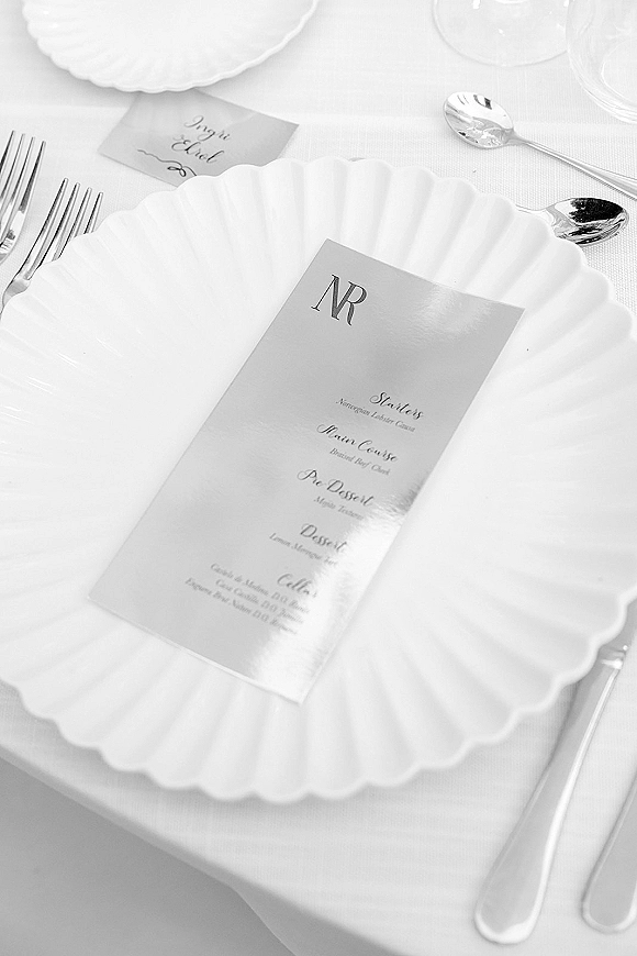Wedding place setting with wedding menu card on a scalloped plate, place card, silverware and wine glass on a white linen table