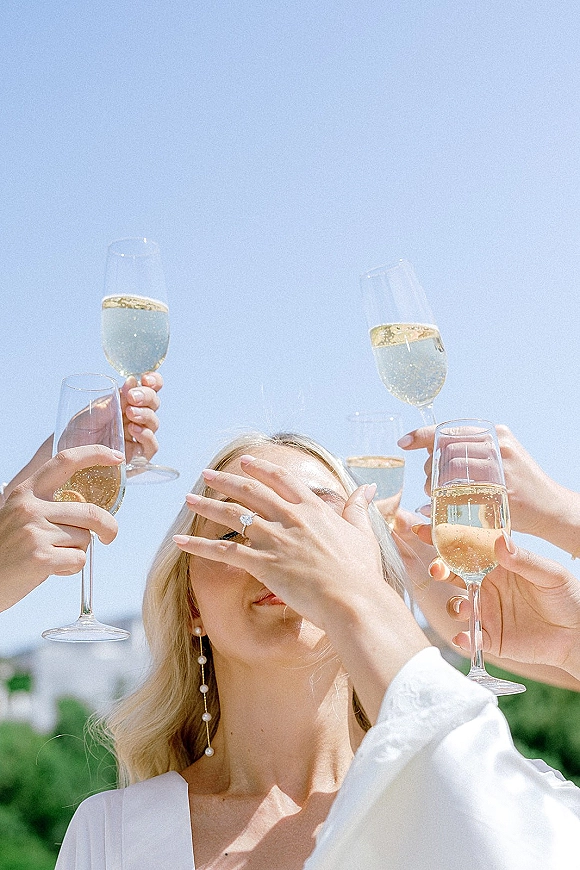 Wedding toast as hands raise champagne flutes of sparkling wine, engagement ring gleaming, pearl drop earrings against blue sky greenery backdrop