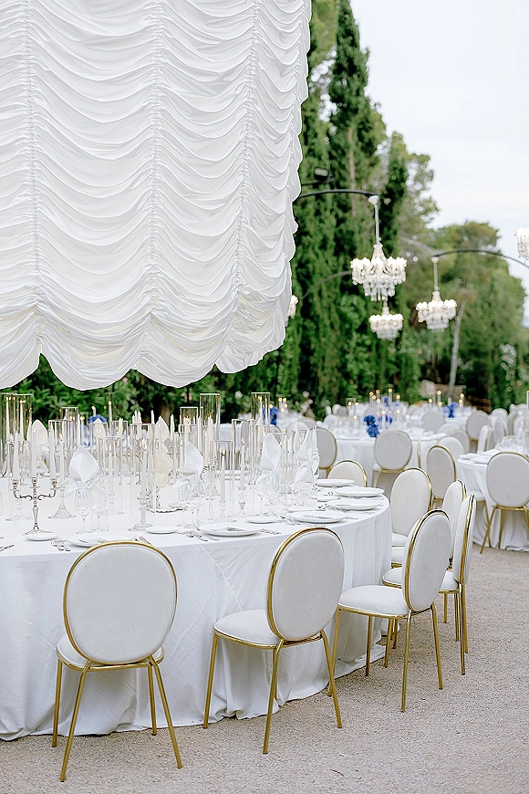 Reception tablescape with white linens, taper candles in glass cylinders, blue floral centerpieces, and chandeliers under a draped canopy outdoors