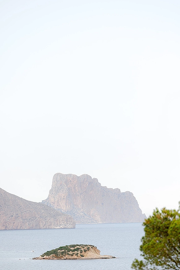 Coastal landscape with ocean cliff view, calm sea and a rocky island below distant cliffs under a pale sky with shrubs in foreground