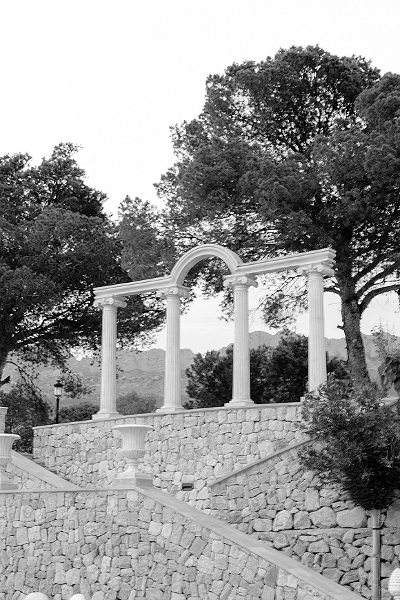 Outdoor wedding venue with classical columns framing an archway and stone steps, set against trees, mountains, and open sky
