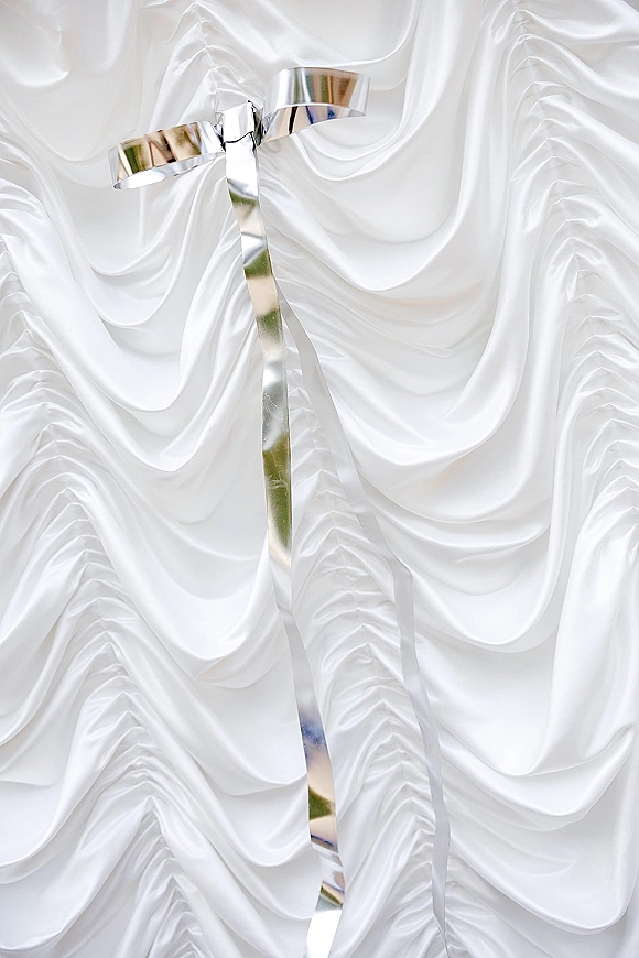 Wedding ribbon with silver wedding ribbon bow and long tails laid on softly draped white satin fabric backdrop for minimal styling detail