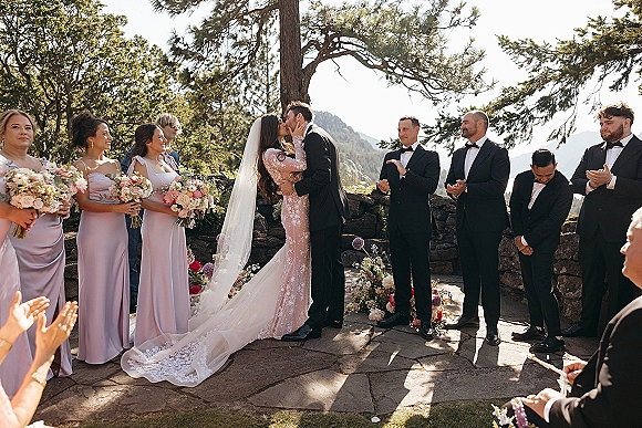 Ceremony kiss moment as bride and groom kiss with a long veil train, wedding party lined up on a stone terrace with mountains behind