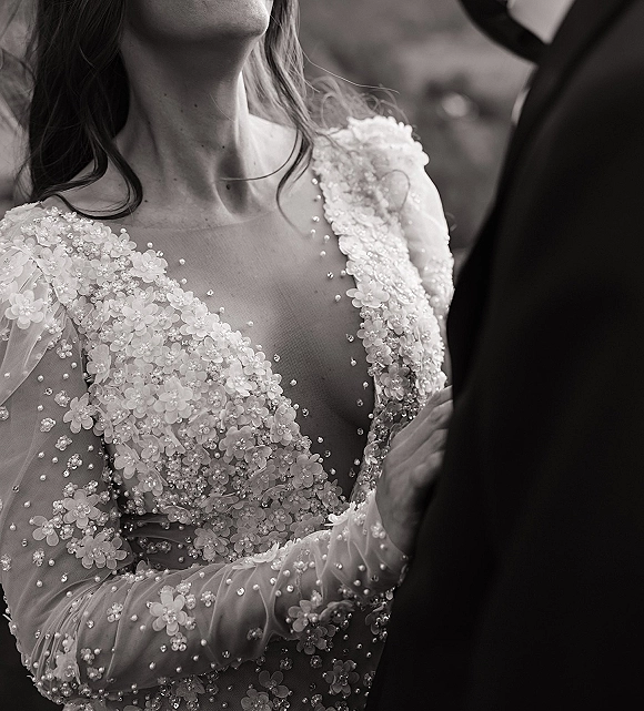 Wedding dress close-up showing an illusion neckline with 3D floral appliqués and beading, with the groom’s suit jacket behind outdoors