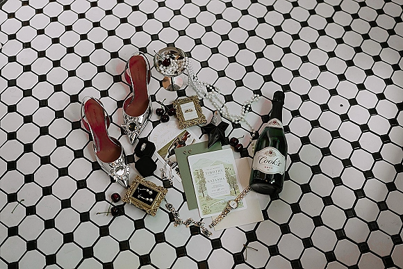 Wedding flat lay with wedding invitation suite, rhinestone bridal heels, pearl jewelry, rings, and champagne on black and white hex tile floor