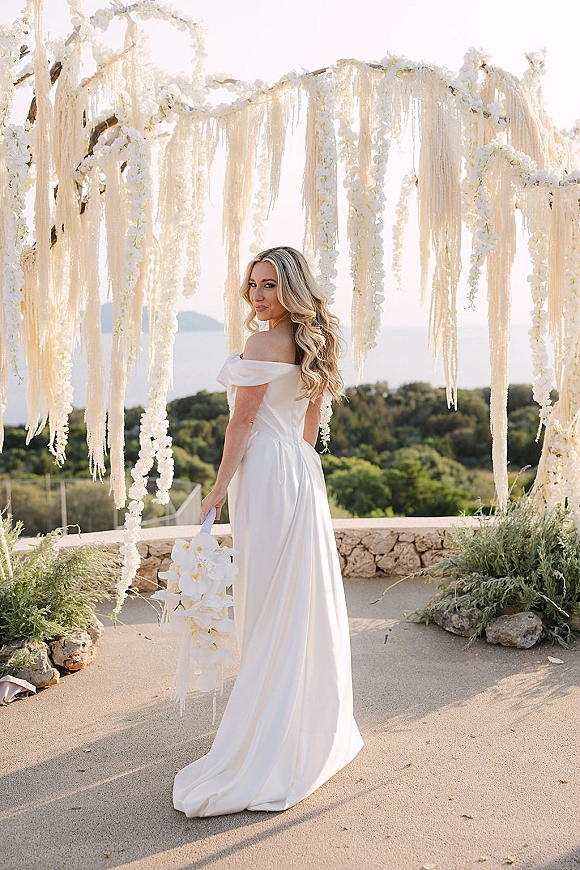 Bridal portrait of a bride looking back in an off-shoulder satin gown holding a white orchid bouquet on an ocean-view terrace under a floral arch