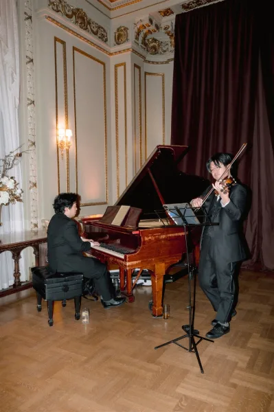 Wedding musicians perform at a grand piano with violin and sheet music, in tuxedos in an ornate gold-trim ballroom with tall curtains