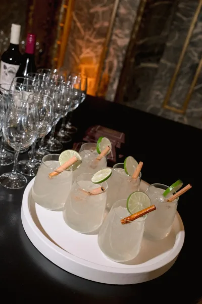 Wedding signature cocktails on a tray with cocktail glasses, lime slices and cinnamon sticks over ice at a warm-lit bar counter, stone wall behind