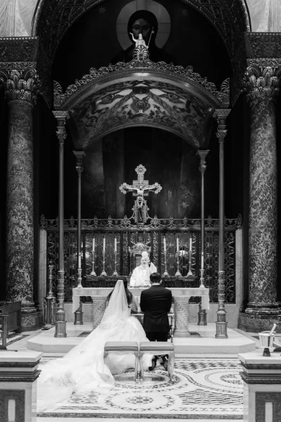 Church ceremony moment in a catholic wedding ceremony as bride and groom kneel at the altar rail with candles, crucifix, and priest under ornate arches