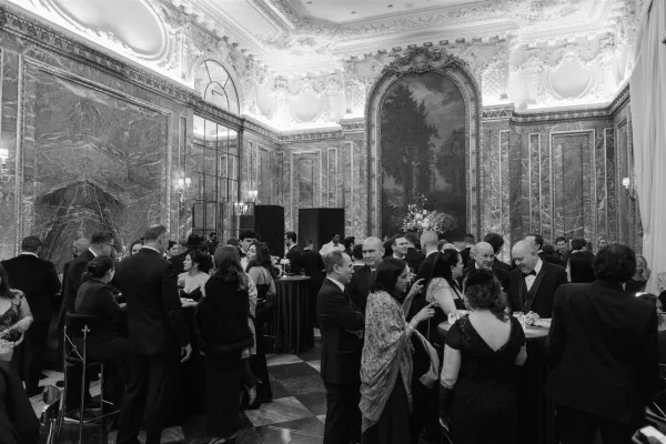 Wedding cocktail hour with wedding guests mingling around cocktail tables, dressed in black tie, beneath chandeliers in an ornate marble ballroom