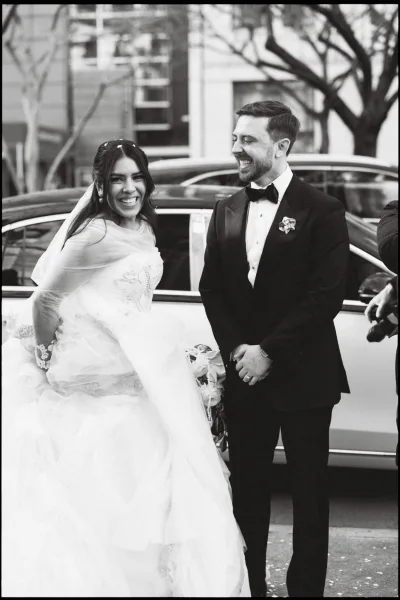 Couple portrait in a black and white wedding photo, bride and groom laughing on a tree-lined street as she holds a bouquet by the car