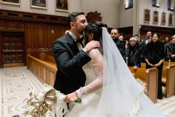 Ceremony moment as groom kisses bride’s forehead while she holds calla lily bouquet, veil trailing in a candlelit church aisle with pew guests
