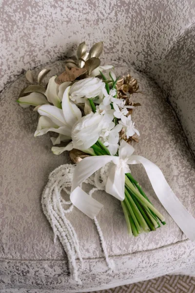 Bridal bouquet with white lily blooms, greenery stems, and a ribbon bow resting on an upholstered chair with textured fabric backdrop