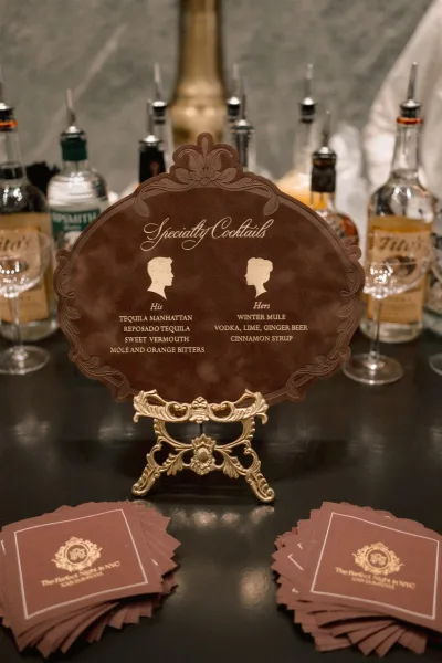 Signature cocktail sign with his and hers cocktails sign on an ornate gold easel beside liquor bottles and recipe cards at the bar counter