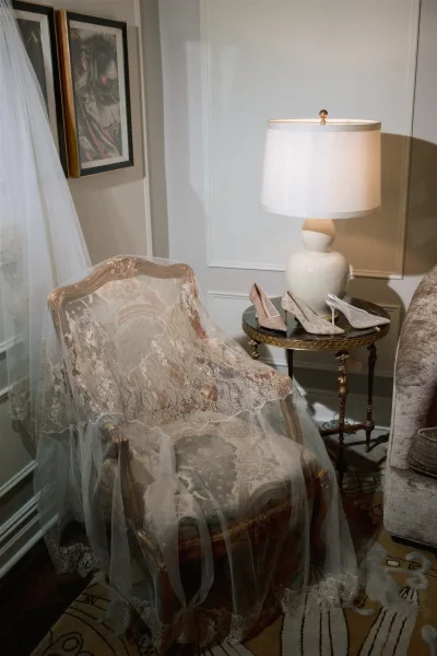 Bridal veil detail with lace wedding veil draped over a vintage armchair beside white heels, framed art, and a paneled living room corner