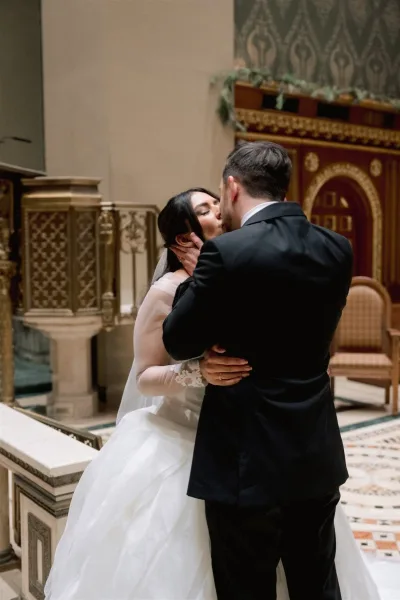 Wedding kiss as bride in long sleeve dress and veil kisses groom in suit at an indoor ceremony by an ornate altar