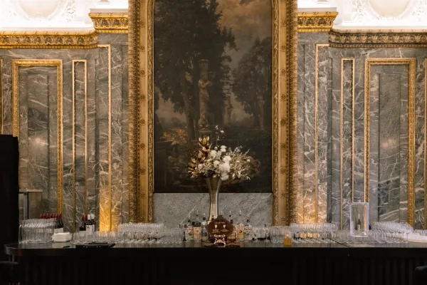 Wedding bar setup with cocktail bar sign, liquor bottles and glassware on a black counter, accented by tall florals against gilded marble walls