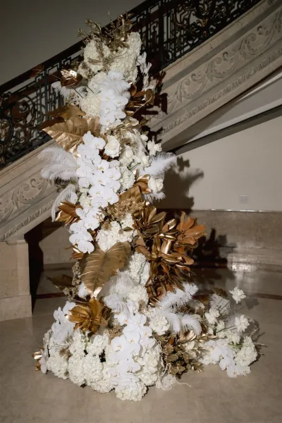 Wedding floral installation of white orchids and roses with gold leaves and pampas grass cascading beside an ornate indoor staircase railing
