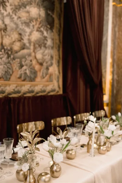 Reception tablescape wedding table decor with white floral centerpieces, gold bud vases and taper candles on a long banquet table before tapestry and dark drapery