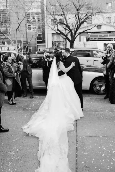 Wedding kiss portrait of bride in ball gown and long veil kissing groom in tuxedo as confetti falls on a city street with guests cheering