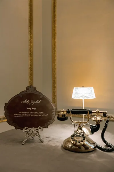 Wedding audio guestbook with a vintage rotary phone, instruction sign, and table lamp on a tabletop against a neutral wall with gold trim molding