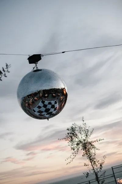 Wedding disco ball hanging by cable wire at sunset, reflecting light against ocean horizon with tree branches and railing behind