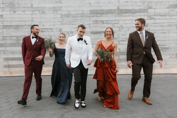 Wedding party portrait of bridesmaids and groomsmen walking with bouquets and boutonnieres by a concrete wall, groom in white jacket