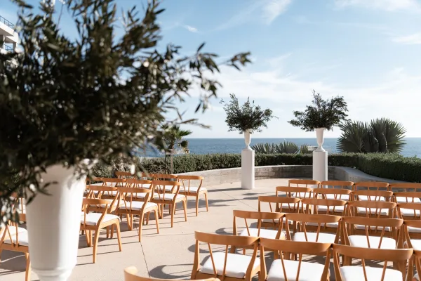 Ceremony setup with outdoor wedding ceremony seating, wood chairs and white cushions facing an ocean-view terrace under blue sky
