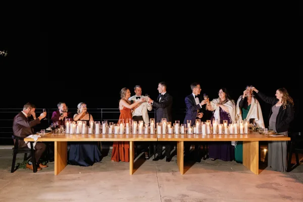 Wedding toast as bridal party raises champagne flutes at a candlelit wooden banquet table on an outdoor patio under the night sky