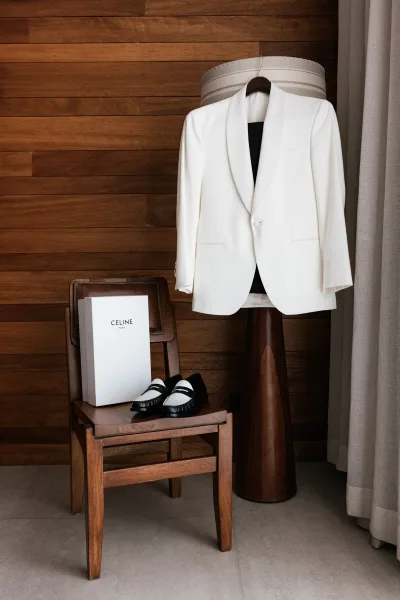 Groom suit details with a white tuxedo jacket on a hanger and leather loafers by a shoebox against a wood-paneled wall