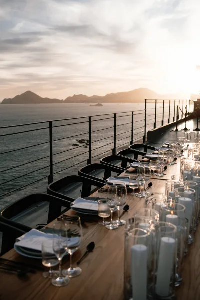 Reception tablescape on a wood dining table with black chairs, menus, wine glasses, and pillar candles on a terrace overlooking ocean at sunset