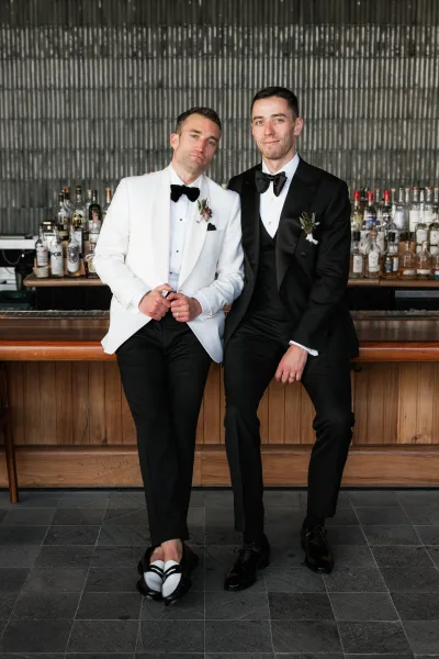 Groom couple portrait of two grooms in tuxedos seated on barstools at a cocktail bar, one in a white dinner jacket with boutonnieres