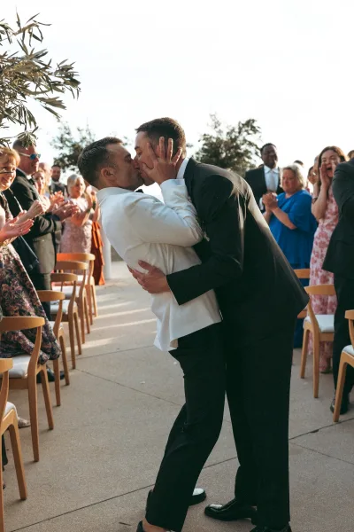 Wedding kiss between two grooms at an outdoor ceremony aisle, one in a white dinner jacket with boutonniere, guests on wooden chairs behind