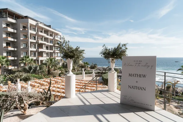 Ceremony setup for an outdoor wedding ceremony with crossback aisle chairs, greenery urns, and a welcome sign on an oceanfront terrace