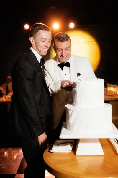 Cake cutting moment as two grooms in black tie tuxedos slice a white two-tier cake on a stand in a dimly lit reception room