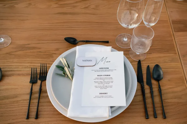 Wedding place setting with wedding menu card on a gray plate, black flatware, white napkin, olive sprig, and clear goblets on a wood table
