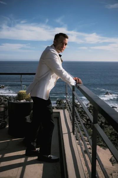 Groom portrait in a white tuxedo jacket with black bow tie and boutonniere, leaning on balcony railing above ocean cliffs under blue sky